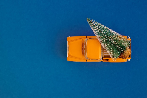 Un'auto gialla in miniatura nella parte posteriore trasporta un albero di Natale su un semplice sfondo blu acquamarina
