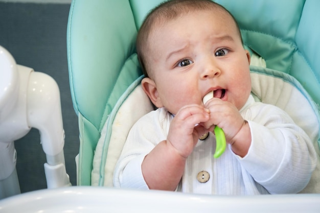 Un bambino affamato sta rosicchiando un cucchiaio di plastica a tavola su un seggiolone Capricci di dentizione gengive pruriginose introduzione di alimenti complementari