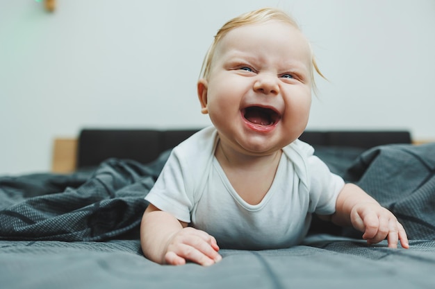 Un bambino allegro con bellissimi occhi grandi è sdraiato sul letto a casa in un bodysuit bianco su lenzuola grigia il bambino è seduto a letto a casa