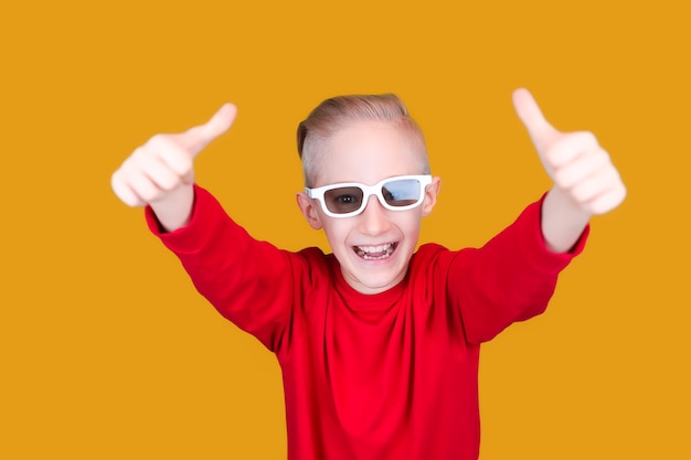 Un bambino allegro in abiti rossi e occhiali mostra un pollice in alto su uno sfondo giallo