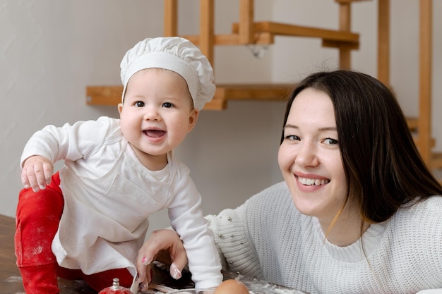 Un bambino asiatico in cappello da chef e grembiule con una giovane madre cucina biscotti allo zenzero di Natale a casa sul tavolo della cucina sorride felicemente e guarda nella telecamera