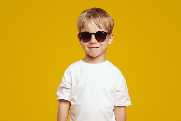 un bambino biondo fiducioso che indossa una maglietta bianca e occhiali da sole freschi sorridendo e guardando la telecamera