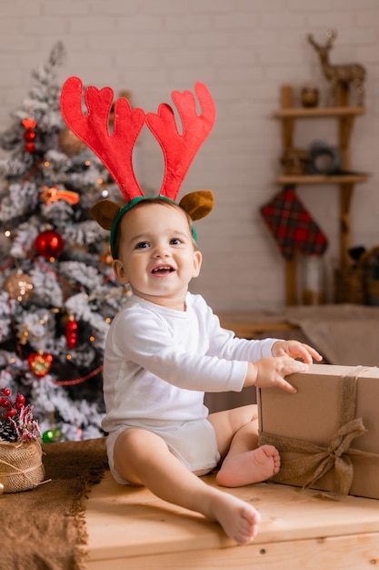 Un bambino carino con un body bianco e corna di renna è seduto con i regali in cucina a casa per Natale il bambino guarda i regali per il nuovo anno