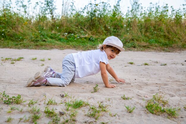 Un bambino carino e felice con un berretto striscia a quattro zampe lungo una strada di campagna nella sabbia in un'estate soleggiata