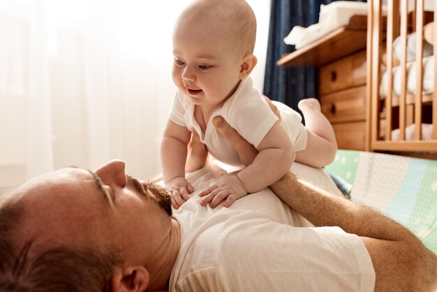 Un bambino carino e gioioso e suo padre in maglietta bianca e jeans giacciono sul pavimento in una stanza luminosa e p