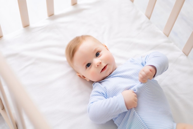 Un bambino carino e sano è sdraiato sulla schiena in una culla su lenzuola bianche a casa in un body blu Il bambino felice guarda nella telecamera sorride