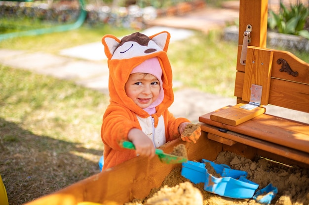 Un bambino carino in tuta arancione gioca nella sabbia nella sandbox all'aperto in una giornata di sole