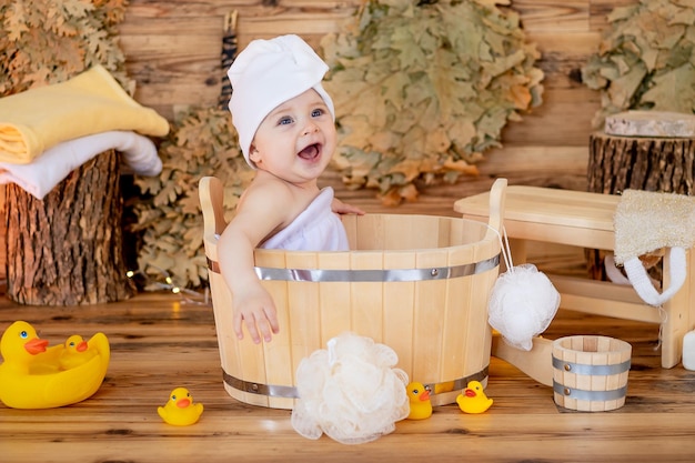 un bambino carino in un asciugamano e un turbante sulla testa dopo il bagno si siede in un bacino in una stanza a vapore in un bagno con un panno e sorride o ride