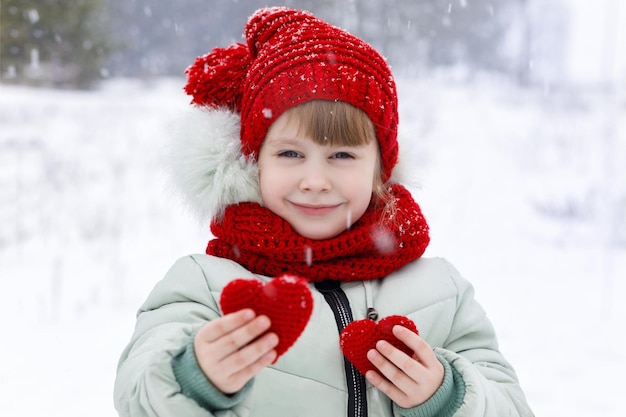 Un bambino carino sorridente sta mostrando due piccoli cuori rossi lavorati a maglia nelle sue mani sullo sfondo del paesaggio invernale innevato