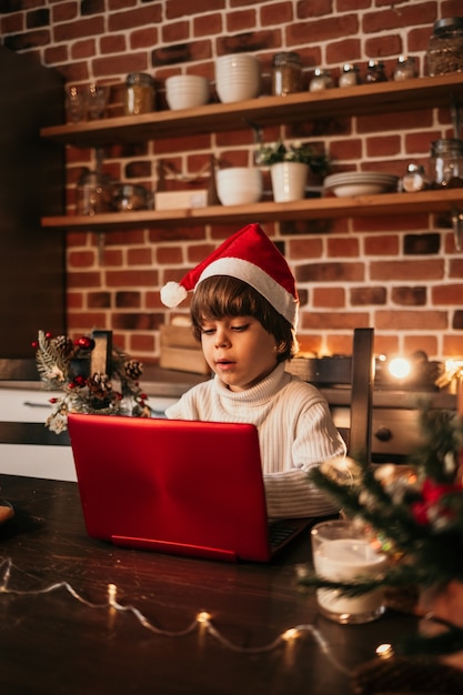 Un bambino caucasico è seduto a un tavolo in cucina e guarda il saluto di Capodanno su un laptop