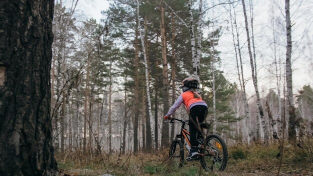 Un bambino caucasico guida su strada in bicicletta nel parco autunnale Bambina che cavalca ciclo arancione nero nella foresta Il bambino va a fare sport in bicicletta Giro in moto motociclista con zaino e casco Mountain bike hard tail