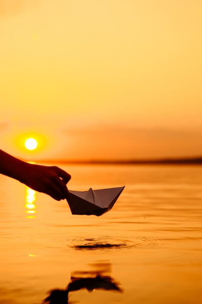 Un bambino che mette una barca di carta in acqua. Tramonto bellissimo. Origami. Fiume. Lago.