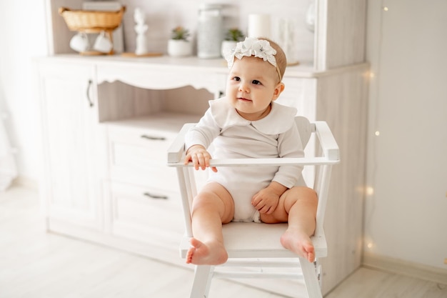 Un bambino con un body di cotone bianco è seduto su un seggiolone in una cucina bianca brillante un piccolo bambino o una bambina sorridente c'è il concetto di cibo per bambini