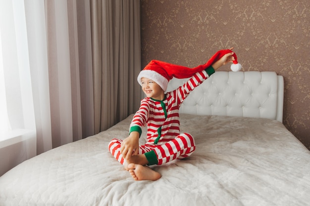 Un bambino con un cappello da Babbo Natale e un pigiama giace su un letto con lenzuola bianche, capodanno, natale