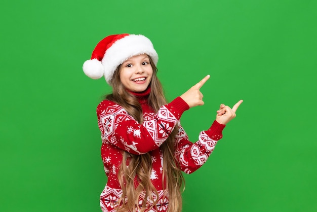 Un bambino con un cappello di Natale Una bambina con un cappello di Babbo Natale e un maglione festivo indica il tuo annuncio su uno sfondo verde isolato