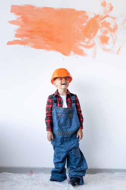 Un bambino con un casco di sicurezza e una tuta sullo sfondo di un muro di cemento bianco con una macchia arancione per lo spazio per il testo Ristrutturazione di una casa o di un appartamento Il ragazzo aiuta i suoi genitori a dipingere