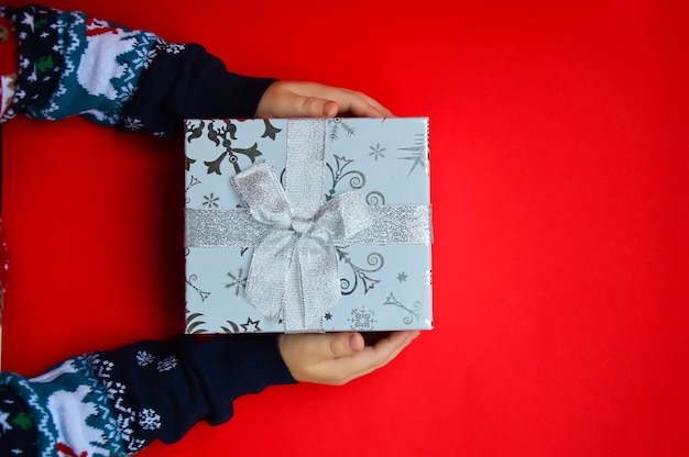 Un bambino con un maglione di Capodanno tiene una scatola regalo di Natale su uno sfondo rosso