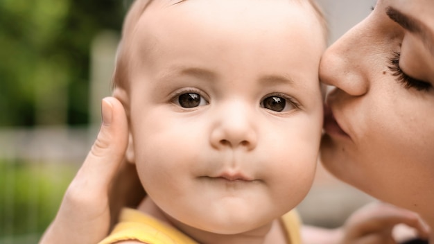 Un bambino con una maglietta gialla che guarda nella telecamera tra le braccia della mamma. La mamma sta baciando suo figlio