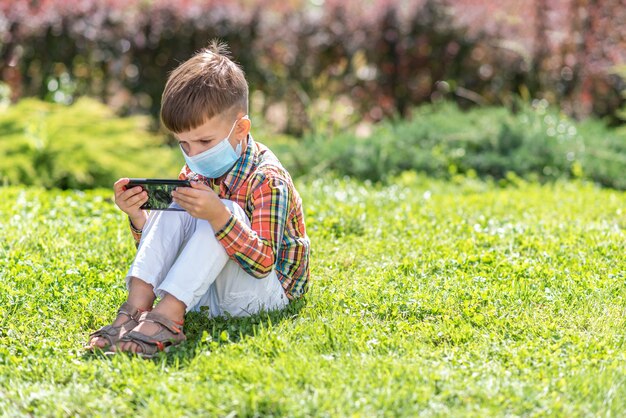 Un bambino con una maschera medica si siede sull'erba e guarda nel telefono