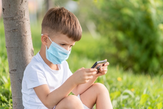 Un bambino con una maschera medica si siede sull'erba e guarda nel telefono