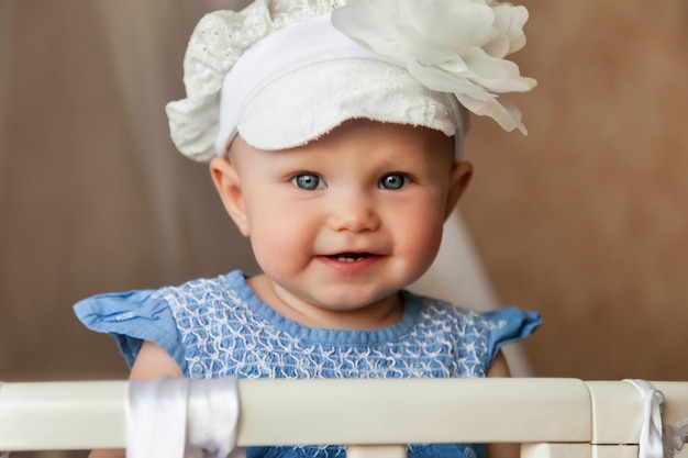 Un bambino dagli occhi azzurri di un anno con un cappello alla moda guarda la telecamera dal presepe. Bambino piccolo e carino con un sorriso seduto nella stanza dei bambini e mamma in attesa. Concetto di corretta educazione e infanzia