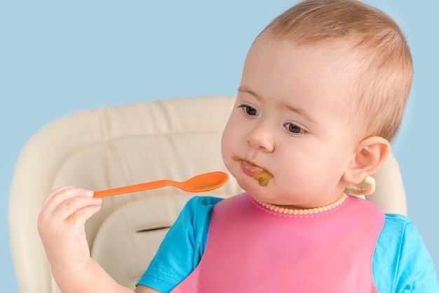 Un bambino di 12-17 mesi con un bavaglino rosa mangia broccoli schiacciati con un cucchiaio mentre è seduto su una sedia. Avvicinamento