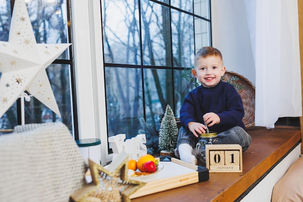 Un bambino di 2 anni si siede sul davanzale della finestra nel nuovo anno atmosfera di Capodanno a casa Il bambino sta aspettando il nuovo anno