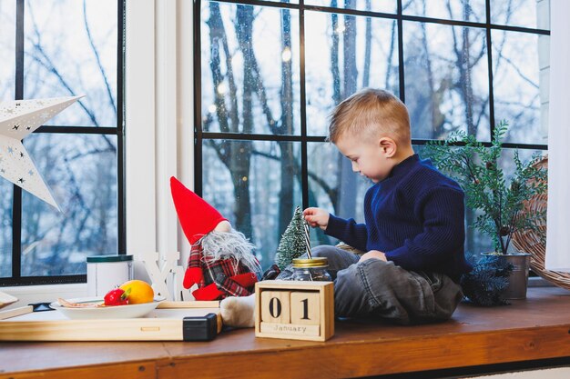 Un bambino di 2 anni si siede sul davanzale della finestra nel nuovo anno atmosfera di Capodanno a casa Il bambino sta aspettando il nuovo anno