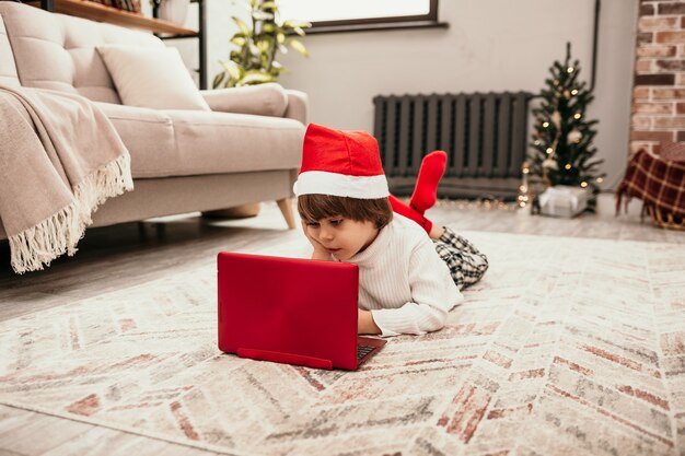 Un bambino di Capodanno con un maglione bianco e un cappello rosso di Natale giace sul tappeto nella stanza e guarda un laptop rosso