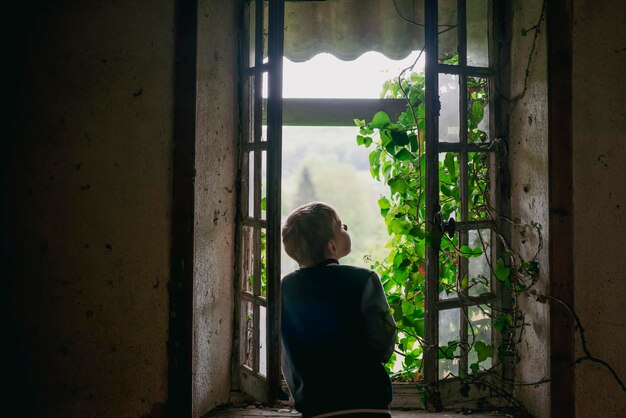 Un bambino di nove anni vicino a una finestra di legno molto vecchia ricoperta di edera in una casa abbandonata