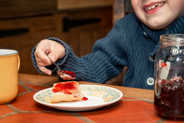 Un bambino di otto anni mangia la marmellata di ciliegie con il pane a tavola in cucina