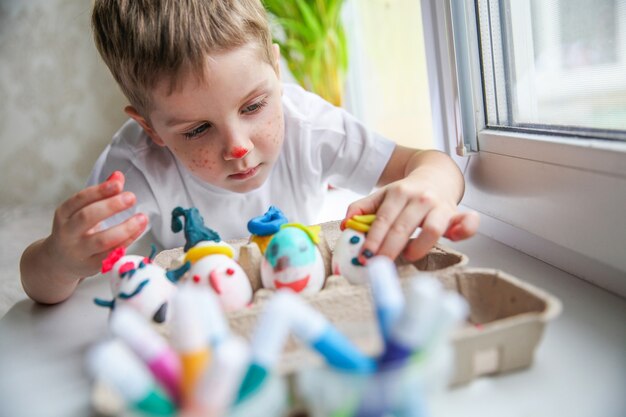 Un bambino di quattro anni con la faccia dipinta sdraiato sul davanzale della finestra mette le uova decorate per la Pasqua in una scatola artigianale