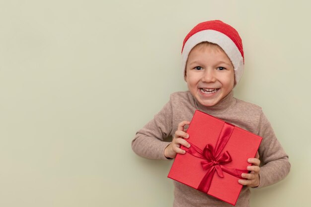 Un bambino divertente in un cappello di Babbo Natale tiene una confezione regalo rossa su uno sfondo chiaro Il concetto di regali di Natale e Capodanno Copia spazio