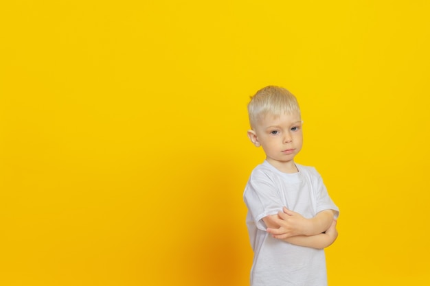 Un bambino è in piedi su un muro giallo, vestito con una maglietta bianca, le braccia incrociate sul petto. Copia luogo.