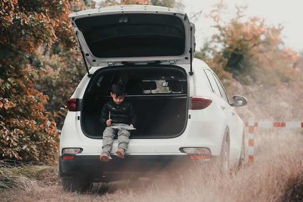 Un bambino è seduto nel bagagliaio di un'auto suv o crossover con guazzo dipinge un'auto in autunno per...