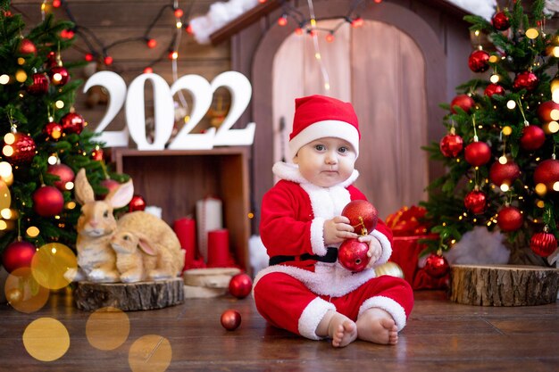 Un bambino è seduto su una slitta sotto gli alberi di Natale. Un grande ritratto. Un bambino in un costume rosso da Babbo Natale con un regalo in una confezione regalo. Buone vacanze, anno nuovo. Periodo natalizio