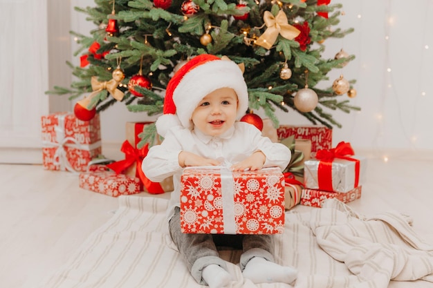 Un bambino felice con un cappello da Babbo Natale si siede sul pavimento vicino all'albero di Natale e tiene in mano una scatola con un regalo.