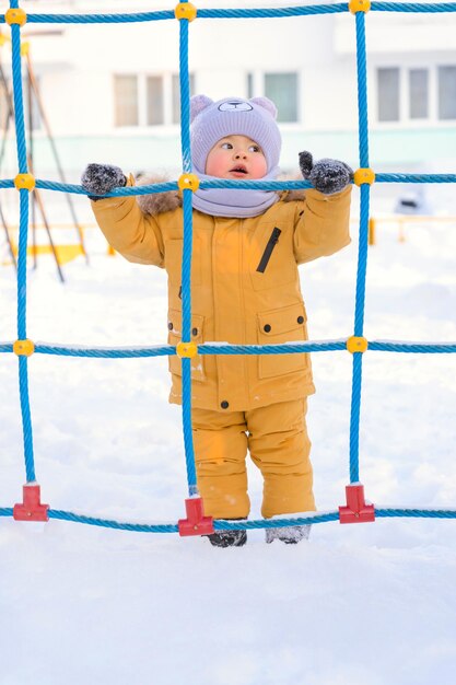 Un bambino felice di 15-25 mesi gioca con una rete di corda in un parco giochi per bambini in inverno