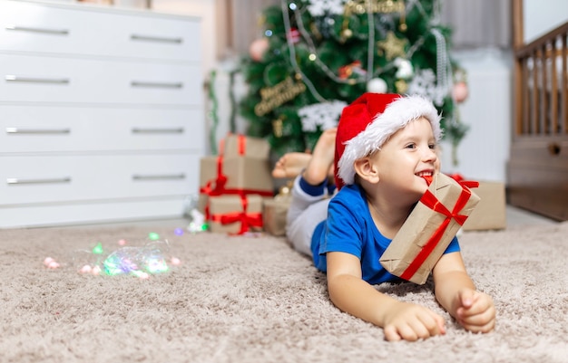 Un bambino felice la mattina di Natale riceve un regalo sdraiato su un tappeto accogliente nella sua stanza in pigiama