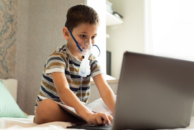 Un bambino impara le lezioni a casa in una maschera d'ossigeno con un nibulizer, un computer portatile e un libro