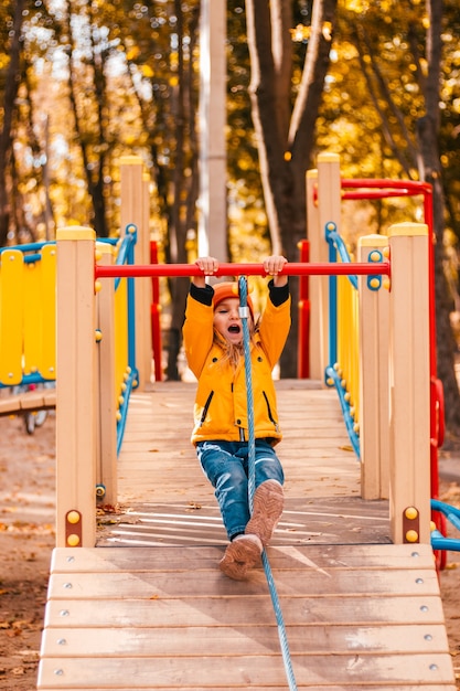 Un bambino in autunno gioca nel parco giochi con una giacca gialla da parco e jeans appesi a una corda