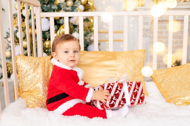 Un bambino in costume da Babbo Natale con un regalo all'albero di Natale con decorazioni dorate è seduto in una culla a casa