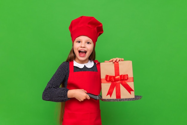 Un bambino in costume da chef tiene una padella per frittelle Una bambina con un grembiule rosso e un cappello tiene un regalo festivo in una padella per una cena su uno sfondo verde isolato