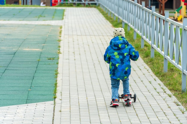 Un bambino in giacca e jeans blu guida uno scooter sul marciapiede nel cortile Il concetto di sicurezza dei bambini per strada Un bambino senza dispositivi di protezione è impegnato in attività ricreative