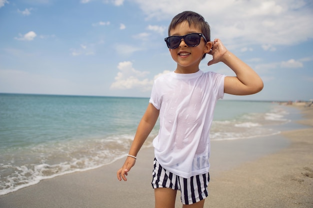 Un bambino in pantaloncini a righe e una maglietta bianca cammina su una spiaggia sabbiosa e indossa occhiali da sole