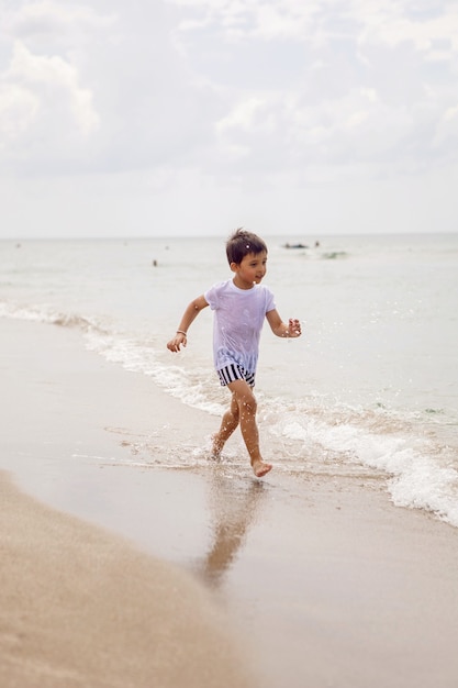 Un bambino in pantaloncini a righe e una maglietta bianca cammina su una spiaggia sabbiosa e indossa occhiali da sole