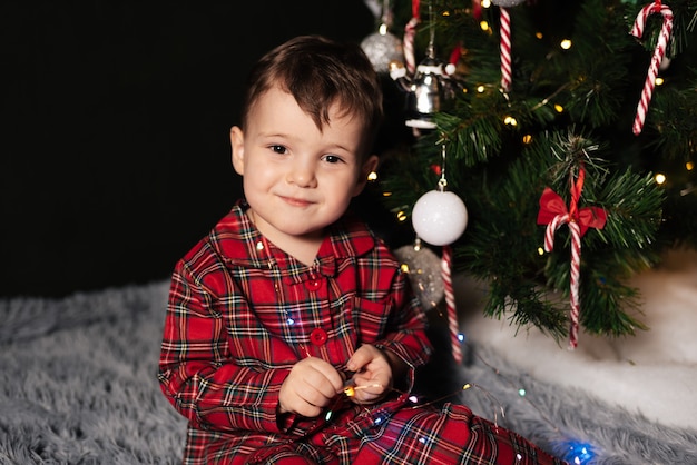 Un bambino in pigiama natalizio vicino a un albero di Natale decorato con decorazioni natalizie