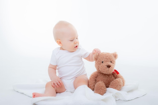 Un bambino in un body bianco si siede con un orsacchiotto su uno sfondo bianco