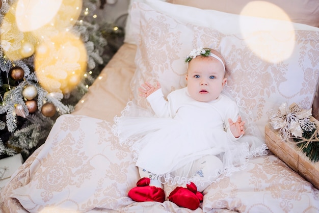 Un bambino in un vestito bianco giace su un divano leggero in ghirlande di luci, un albero di Natale festivo