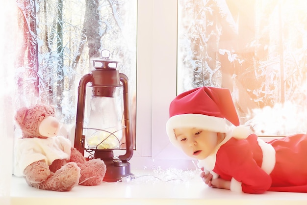 Un bambino nel nuovo anno guarda fuori dalla finestra. I bambini aspettano Babbo Natale. Un bambino vestito da Babbo Natale siede alla finestra.